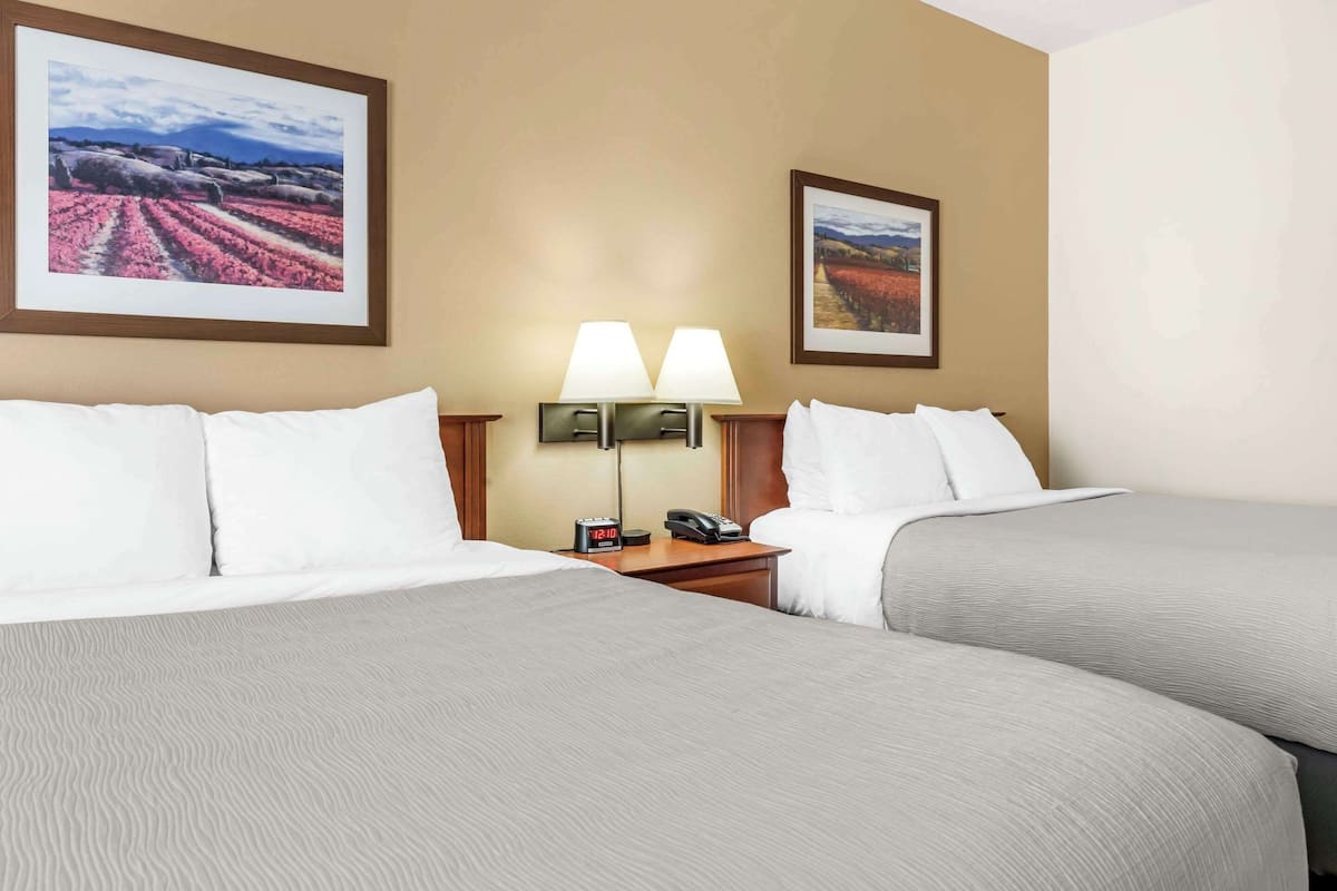 A hotel room with two beds, a nightstand with a lamp, a clock, and two framed pictures on the wall.