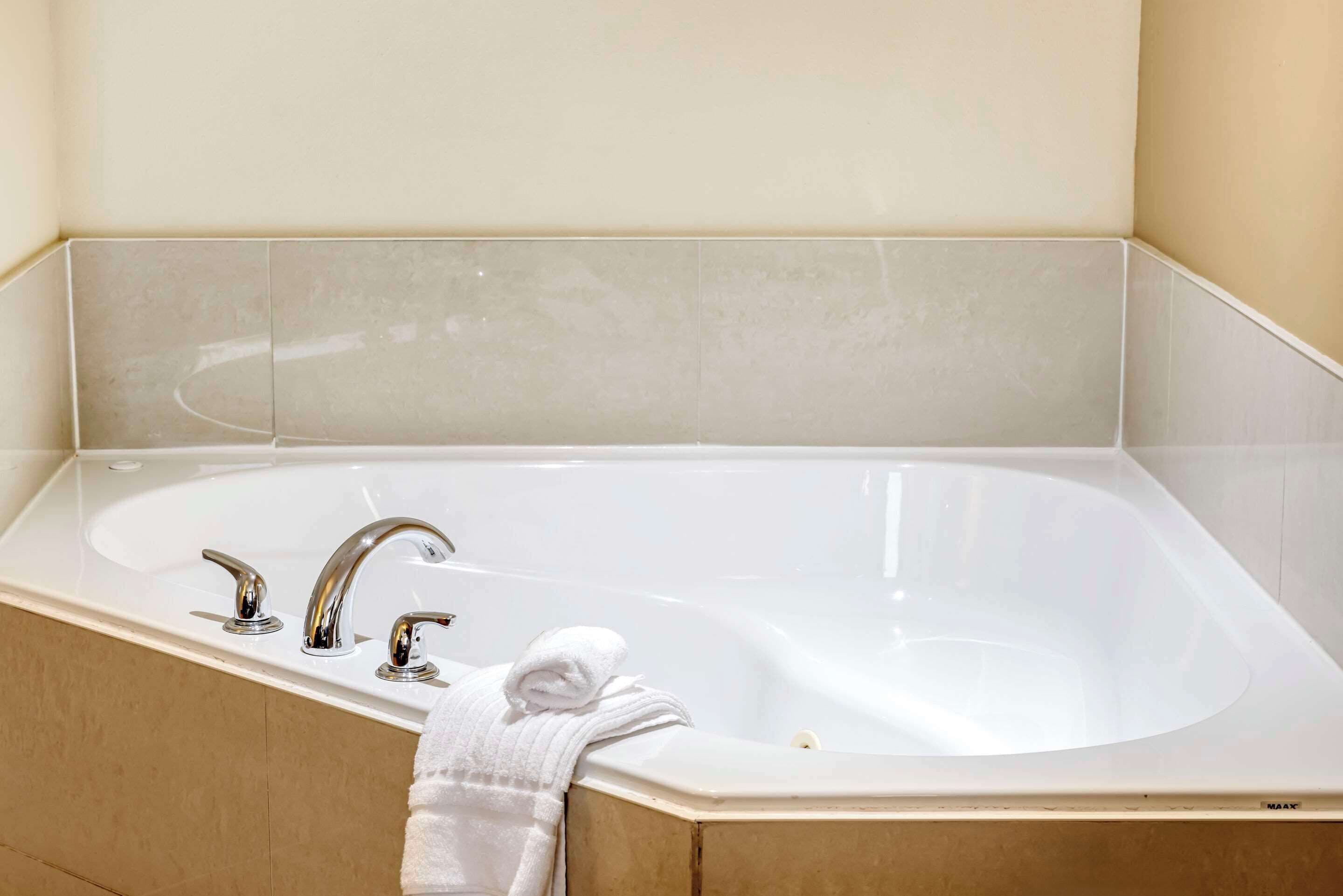 A large, rectangular bathtub with two faucets, a towel, and a beige tiled wall.