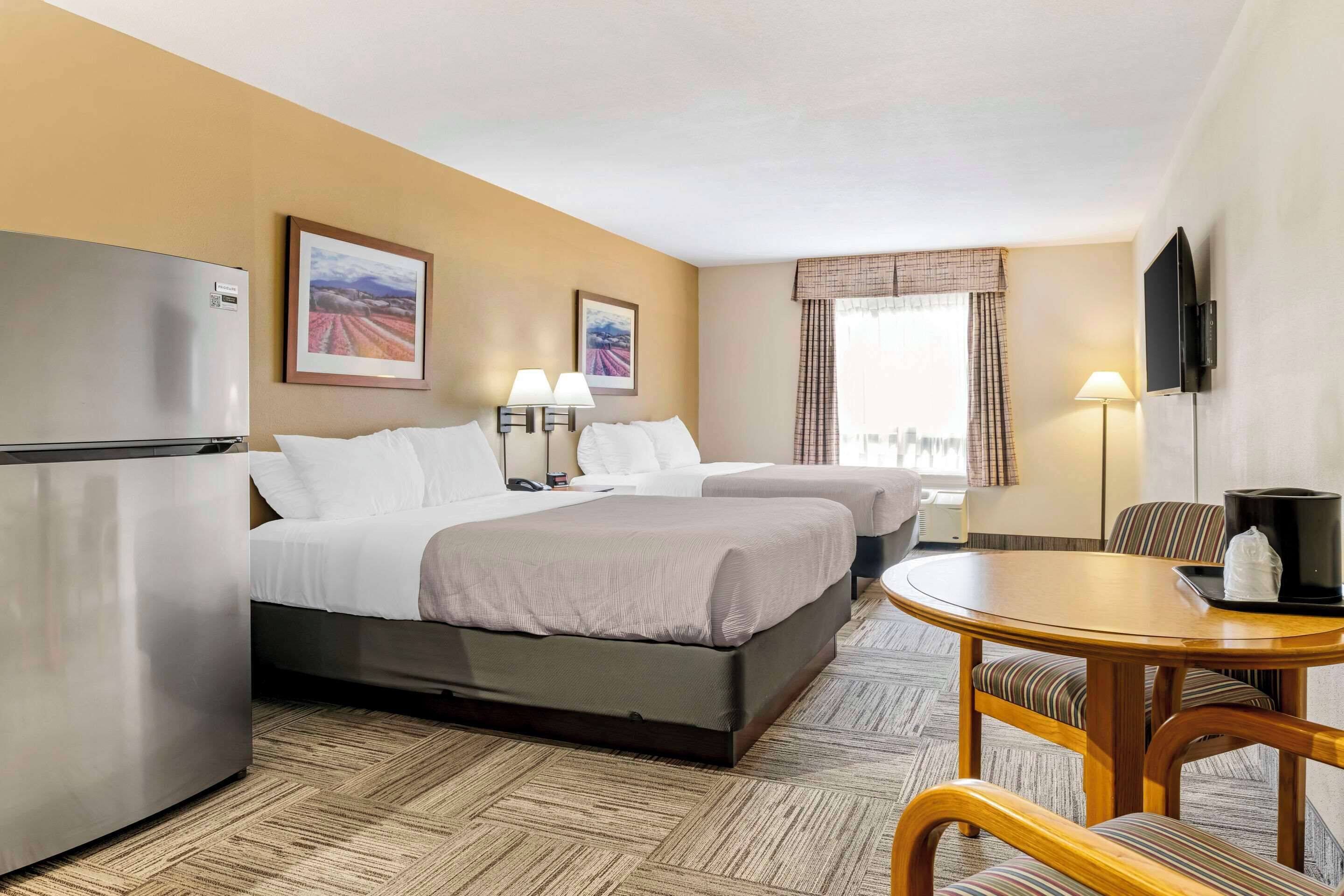 A hotel room with two beds, a refrigerator, a dining table with chairs, a TV, and two wall-mounted lamps.