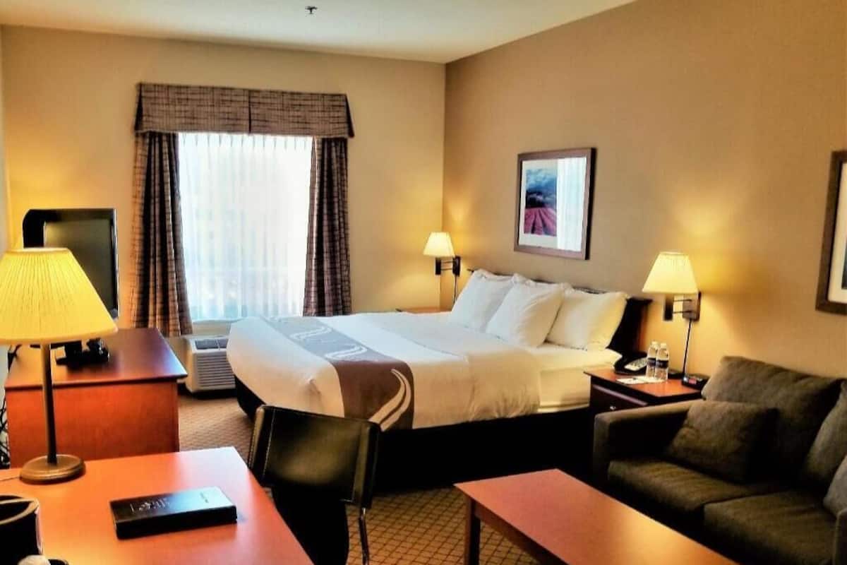 A hotel room with a bed, two bedside tables with lamps, a desk with a chair, a sofa, and a window with curtains.