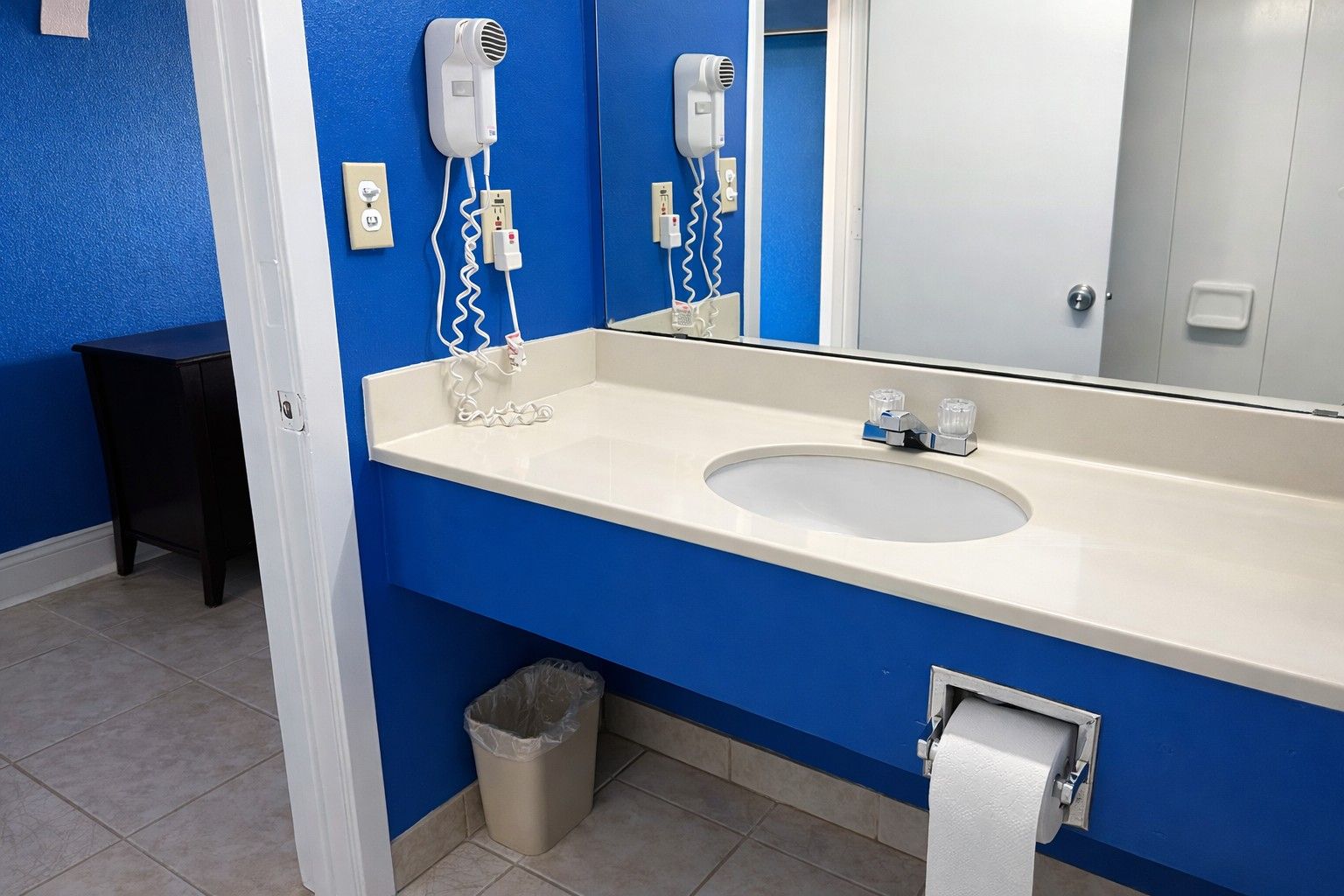 A bathroom with a blue wall, white sink, and a mirror. There are two hair dryers mounted on the wall, a trash can, and a towel dispenser.