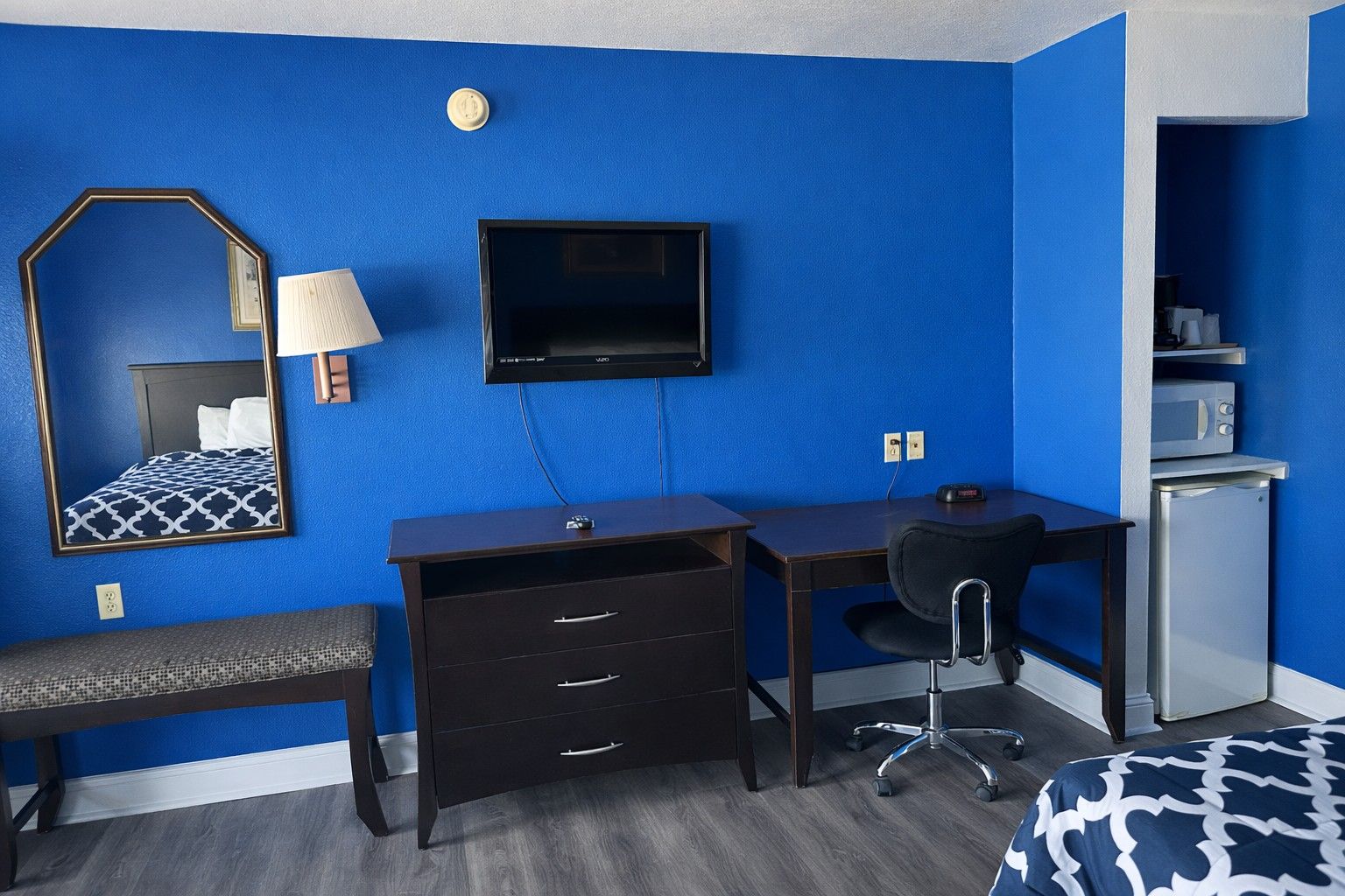 A hotel room with a blue wall, a bed with a patterned bedspread, a desk with a chair, a TV, a small refrigerator, and a mirror with a lamp.