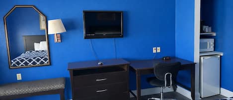Habitación de hotel con pared azul, cama con colcha estampada, escritorio con silla, televisor, refrigerador pequeño y espejo con lámpara.