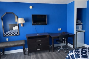 Habitación de hotel con pared azul, cama con colcha estampada, escritorio con silla, televisor, refrigerador pequeño y espejo con lámpara.
