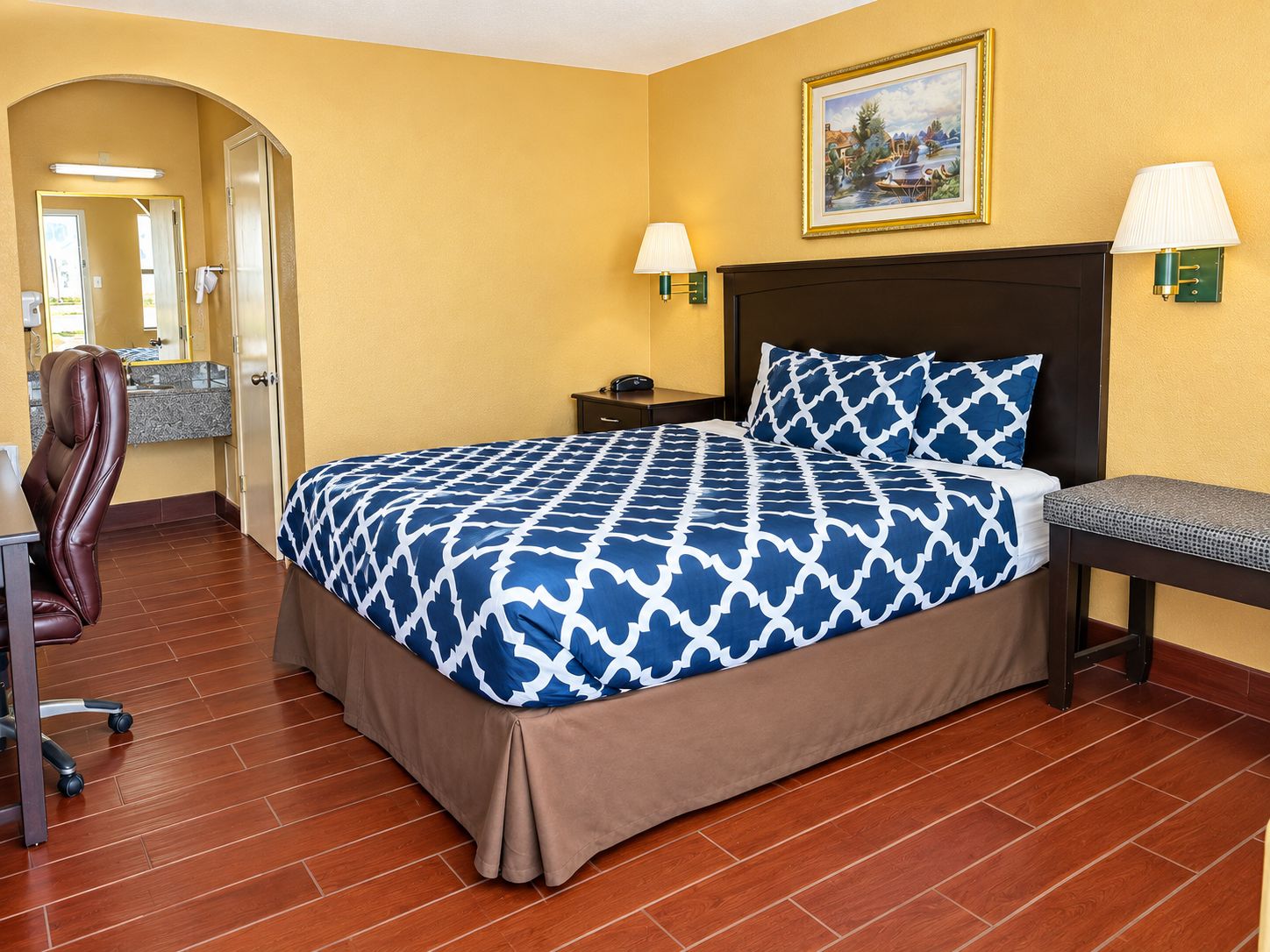 Habitación de hotel con cama, escritorio, silla y baño visible a través de un arco.