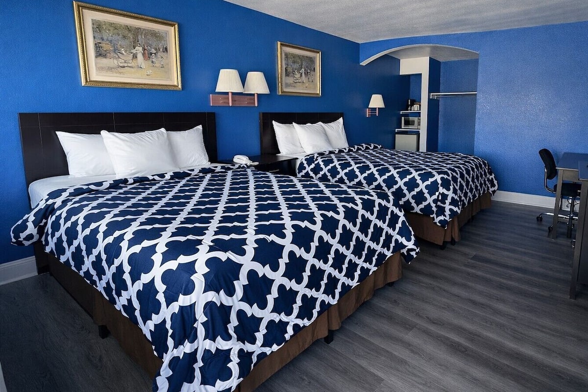 A hotel room with two beds, each with a patterned bedspread, a desk with a chair, and a lamp.