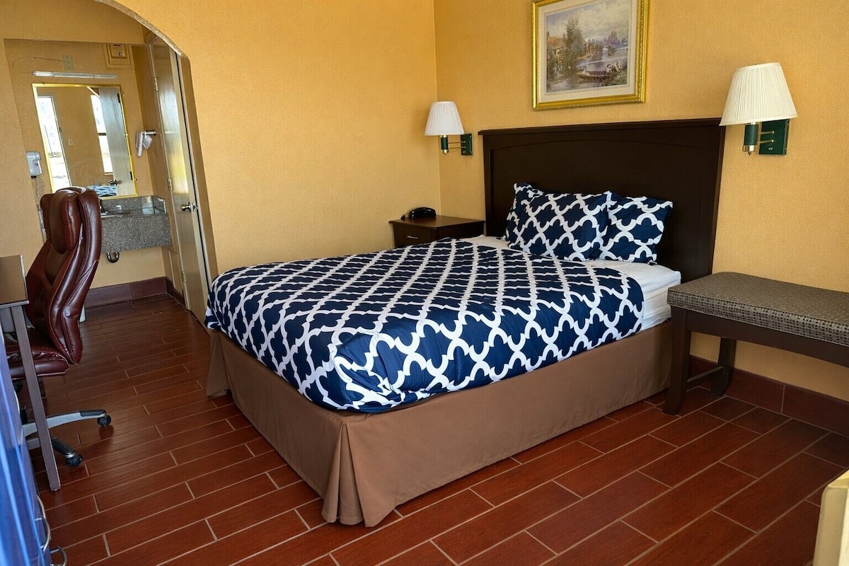 A hotel room with a bed, a bench, a chair, a nightstand, a lamp, and a painting on the wall.