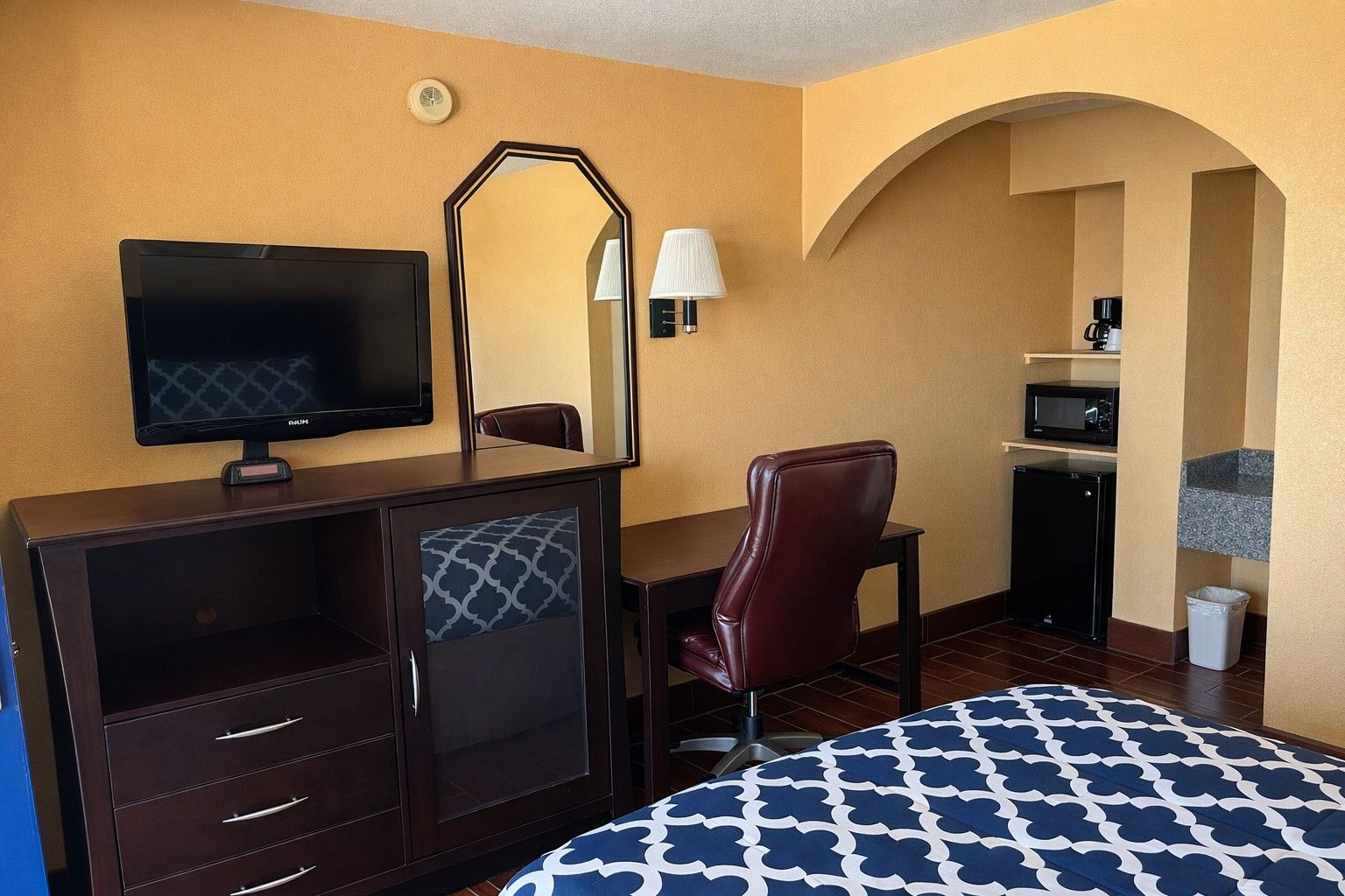 A hotel room with a bed, a desk with a chair, a television, a mirror, a lamp, a microwave, and a small refrigerator.