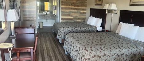 A hotel room with two beds, a desk, a chair, a vanity with a mirror, and a bathroom visible through an open door.
