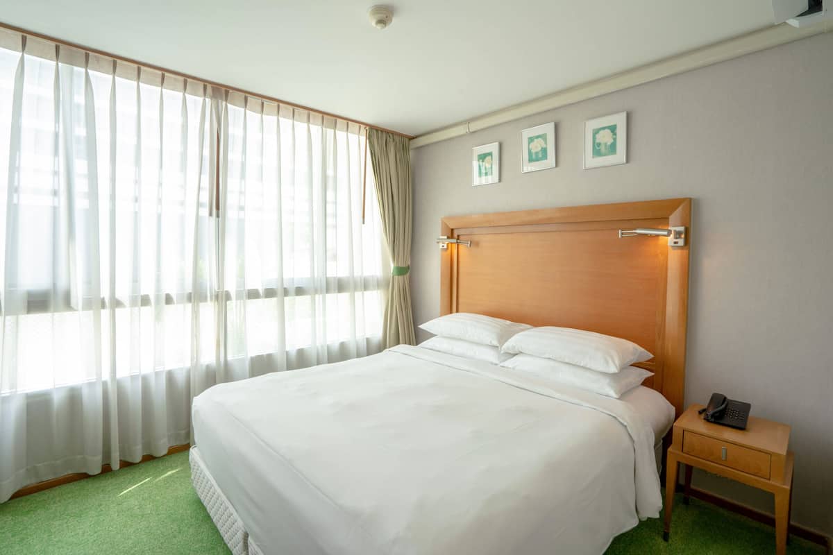 A hotel room with a large bed, a wooden headboard, a bedside table with a phone, and a window with sheer curtains.