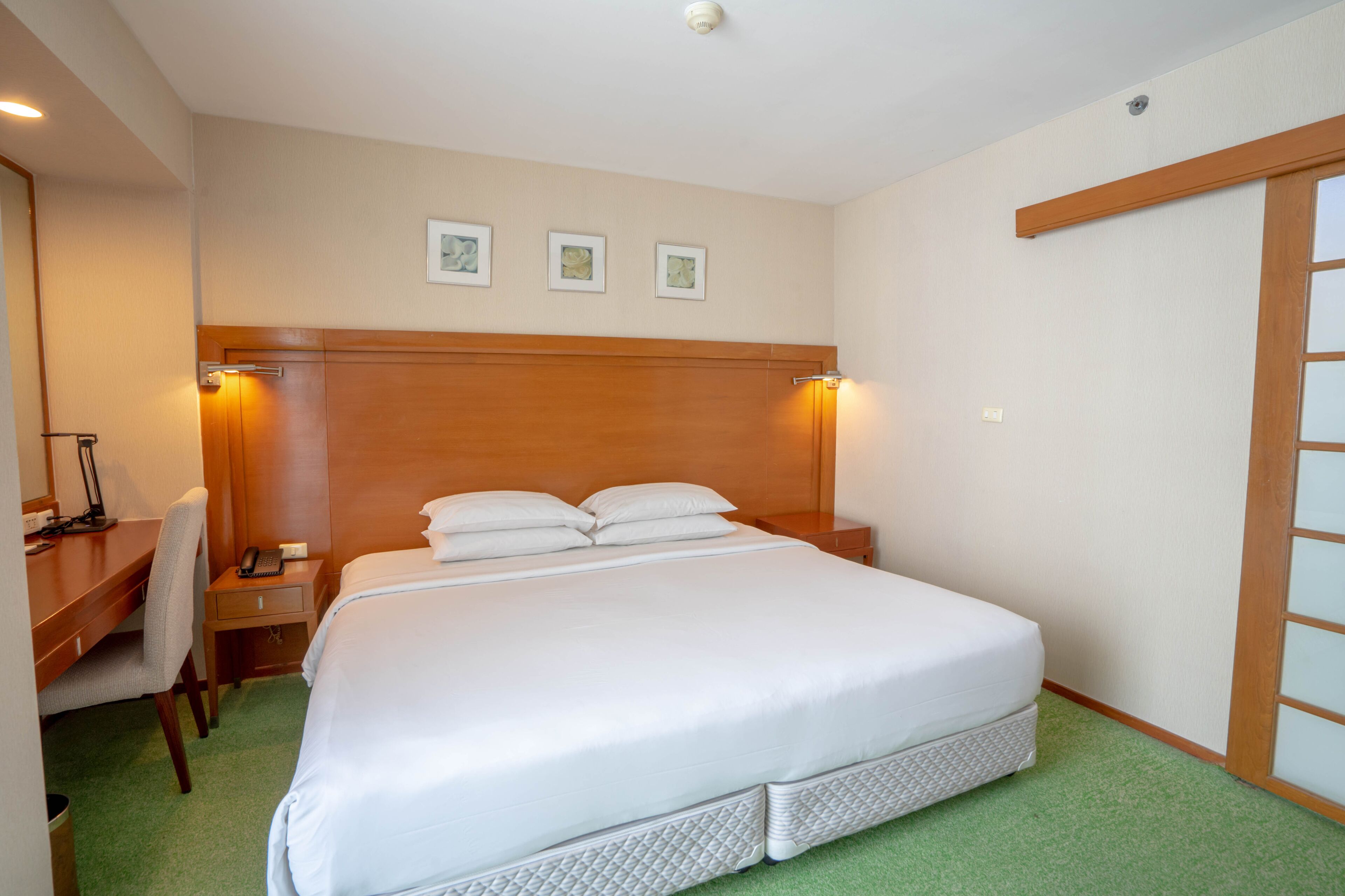 A hotel room with a large bed, a desk with a chair, a nightstand, and a wooden headboard.