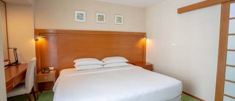 A hotel room with a large bed, a desk with a chair, a nightstand, and a wooden headboard.