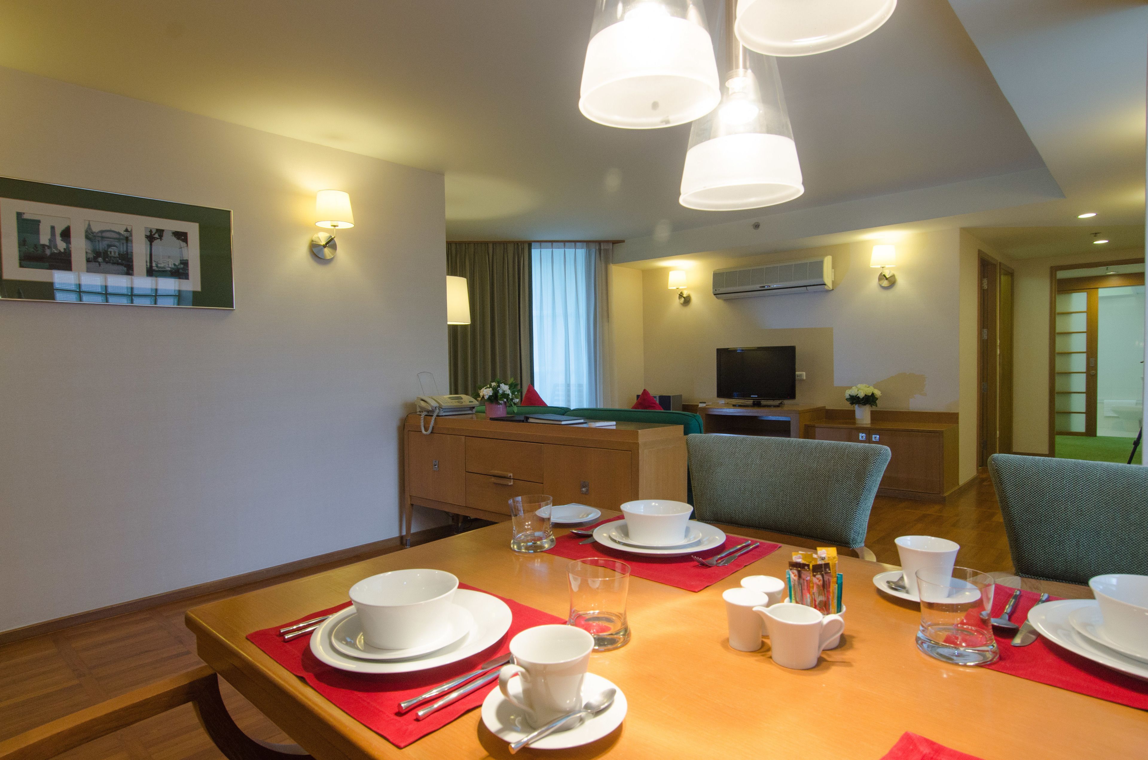 Comedor con mesa de madera dispuesta para cuatro personas, con platos, tazones y tazas blancos. Se observan dos sillas, un televisor y una ventana con cortinas.