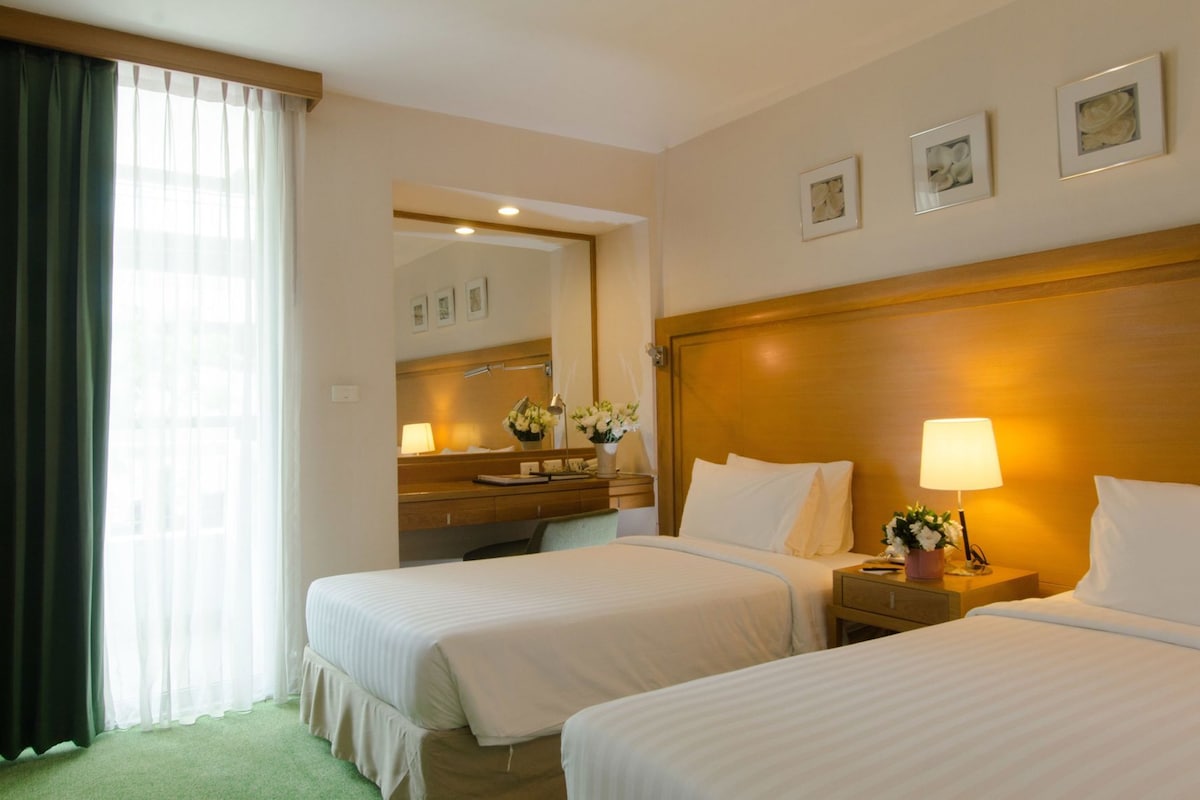 Une chambre d’hôtel avec deux lits, une tête de lit en bois, une table de chevet avec une lampe, un miroir et une fenêtre avec des rideaux.