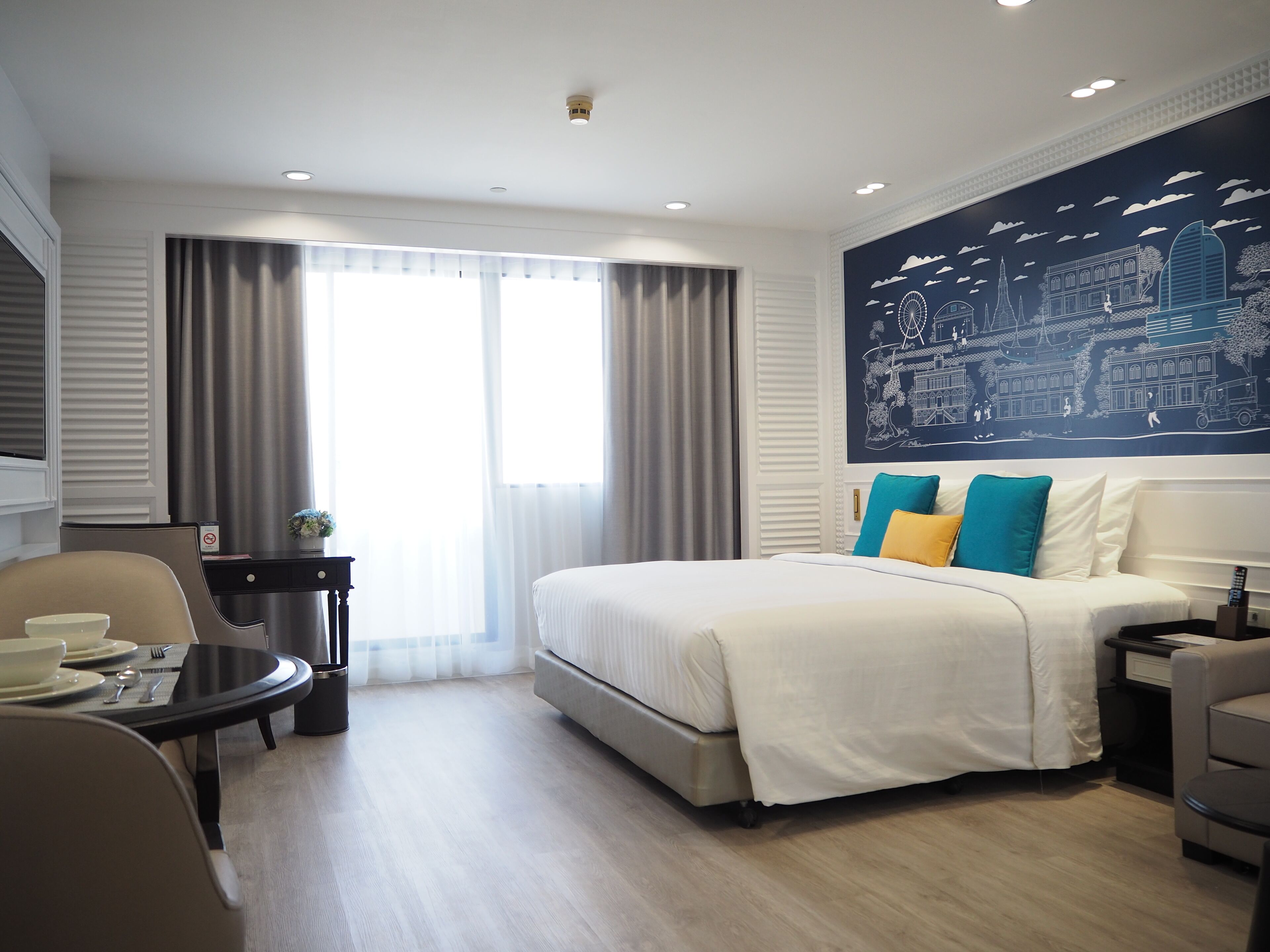 Habitación de hotel con una cama grande, una mesa de comedor con sillas, una mesita auxiliar y una pared con un mural de paisaje urbano.