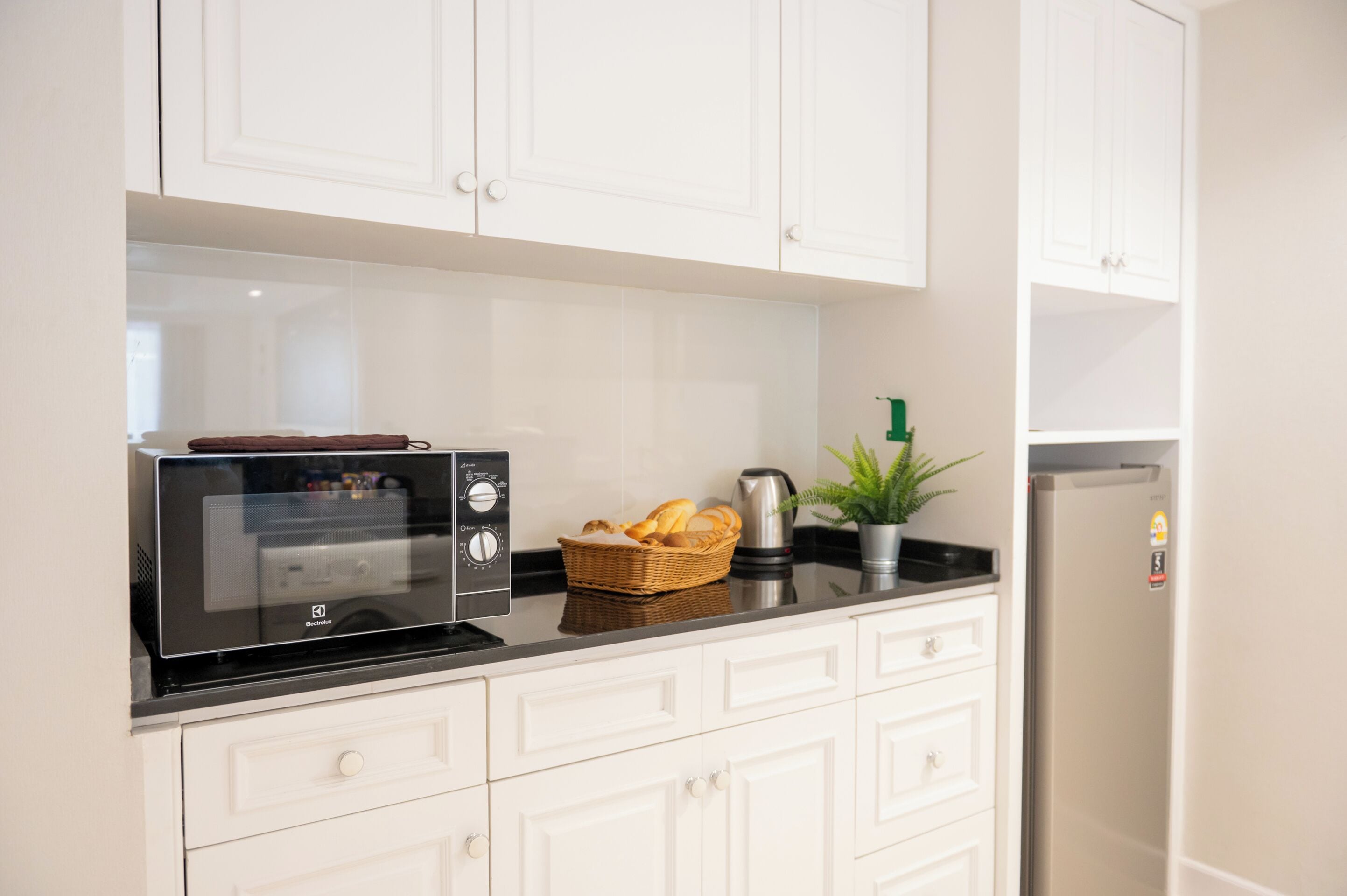 A modern kitchen with white cabinets, a black countertop, a microwave, a basket of bread, a coffee maker, and a refrigerator.