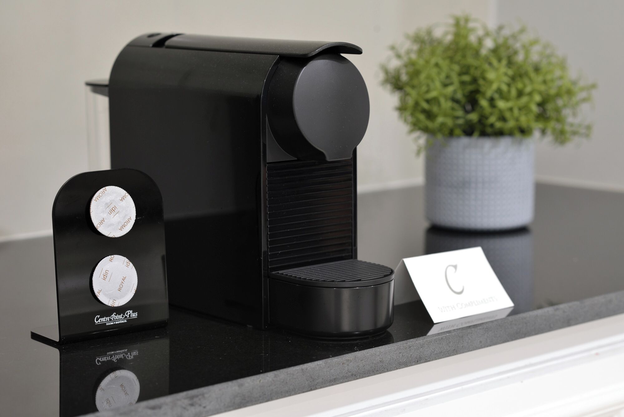 A black coffee maker with a scale on the side, a potted plant, and a card on the counter.