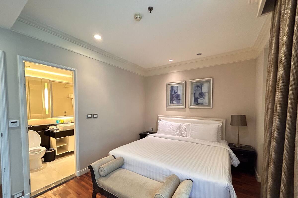 A hotel room with a large bed, a bedside table, a lamp, and a bench.