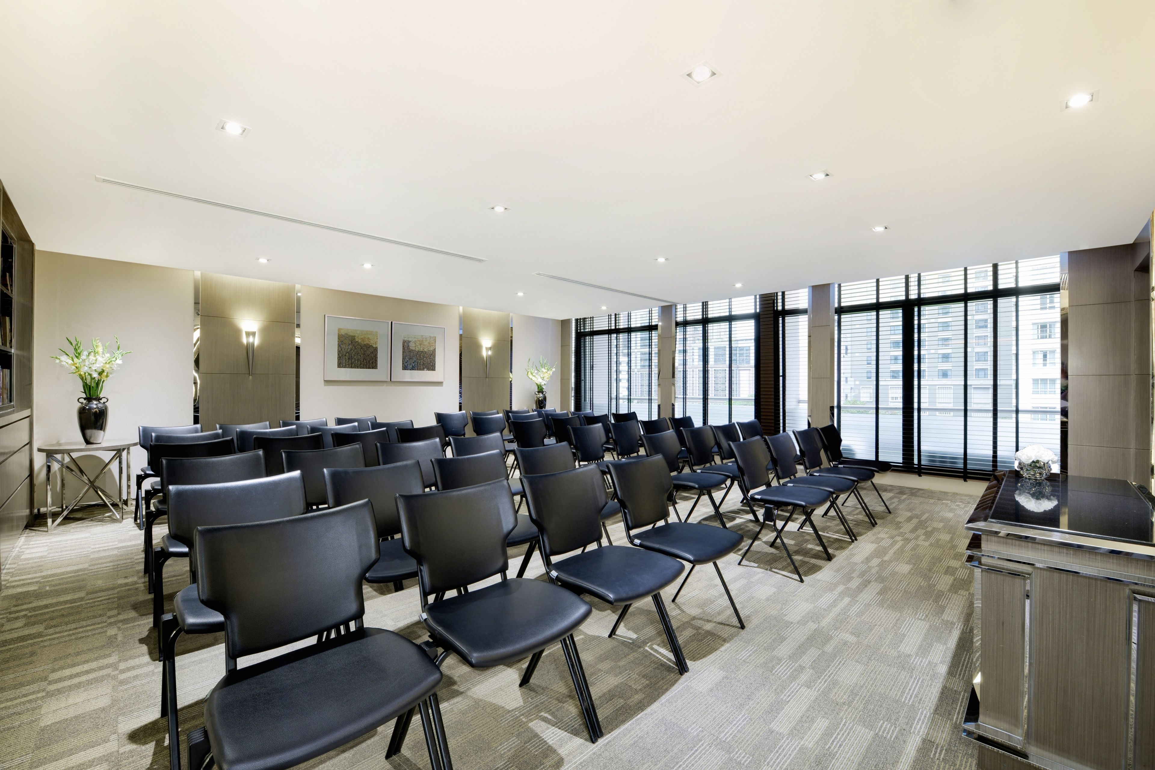Un salón de conferencias con filas de sillas, un atril y grandes ventanales que ofrecen vistas a la ciudad.