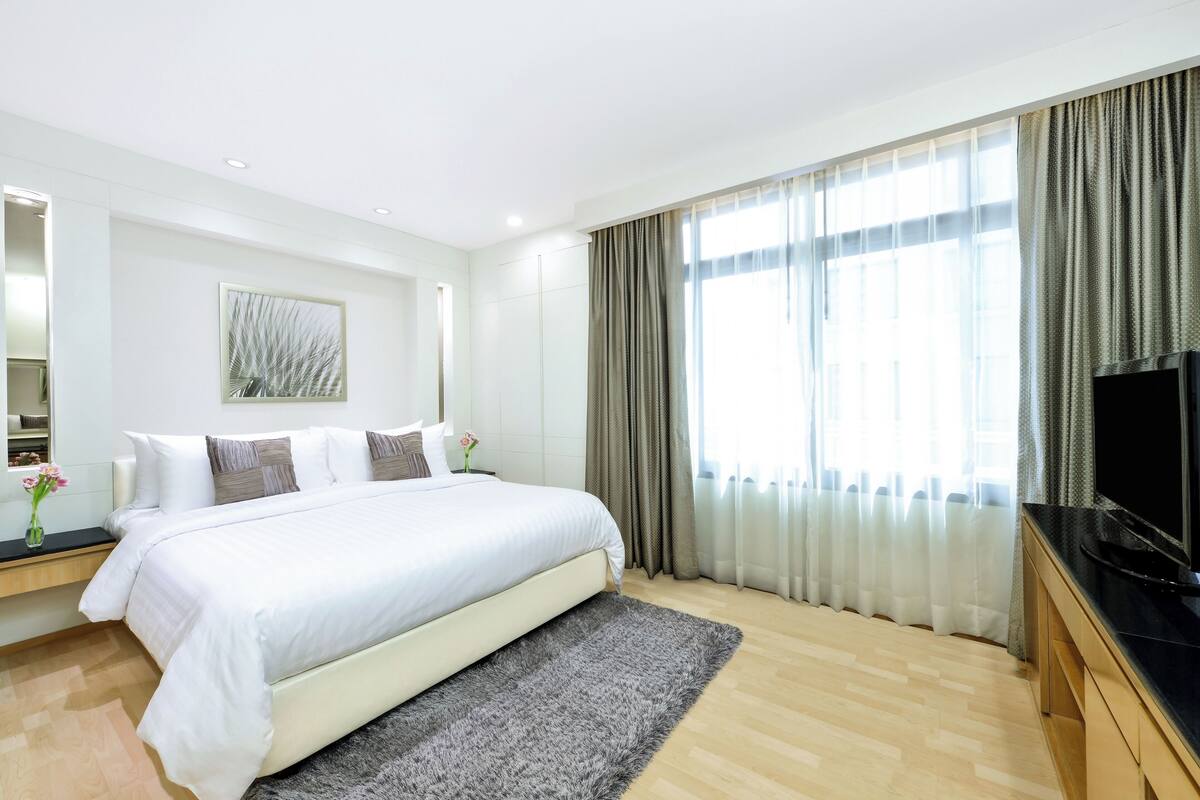 A modern hotel room with a large bed, a flat-screen TV, a wooden floor, and a large window with curtains.