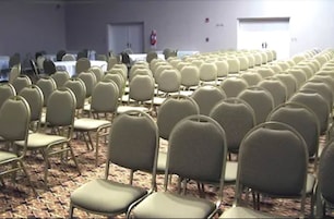 Un salón de conferencias con filas de sillas y mesas, piso alfombrado y un extintor en la pared.