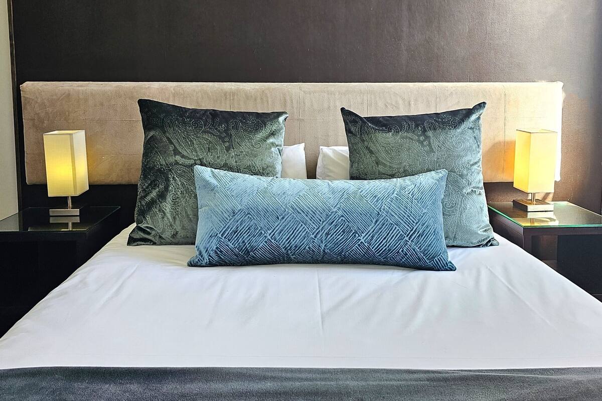 A neatly made bed with a white duvet and three pillows, two with a paisley pattern and one with a geometric design. There are two bedside tables with lamps on either side of the bed.