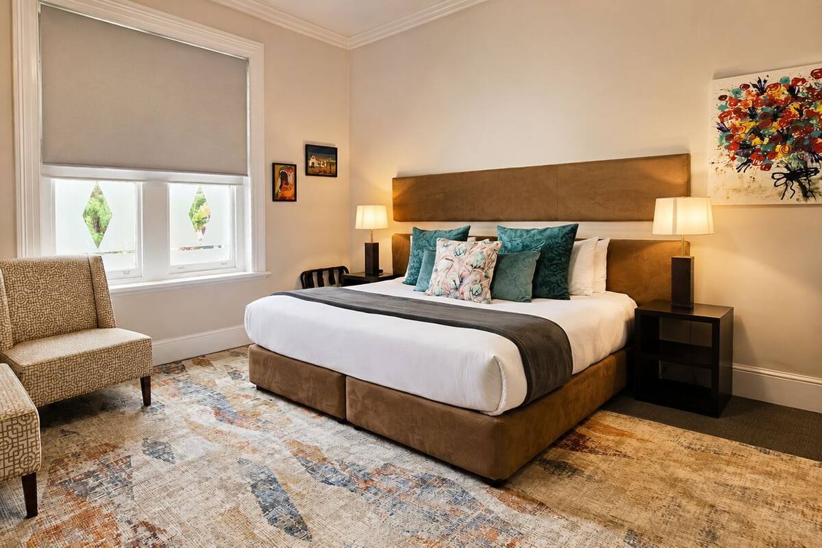 A bedroom with a large bed, two bedside tables, a chair, a window with blinds, and a painting on the wall.