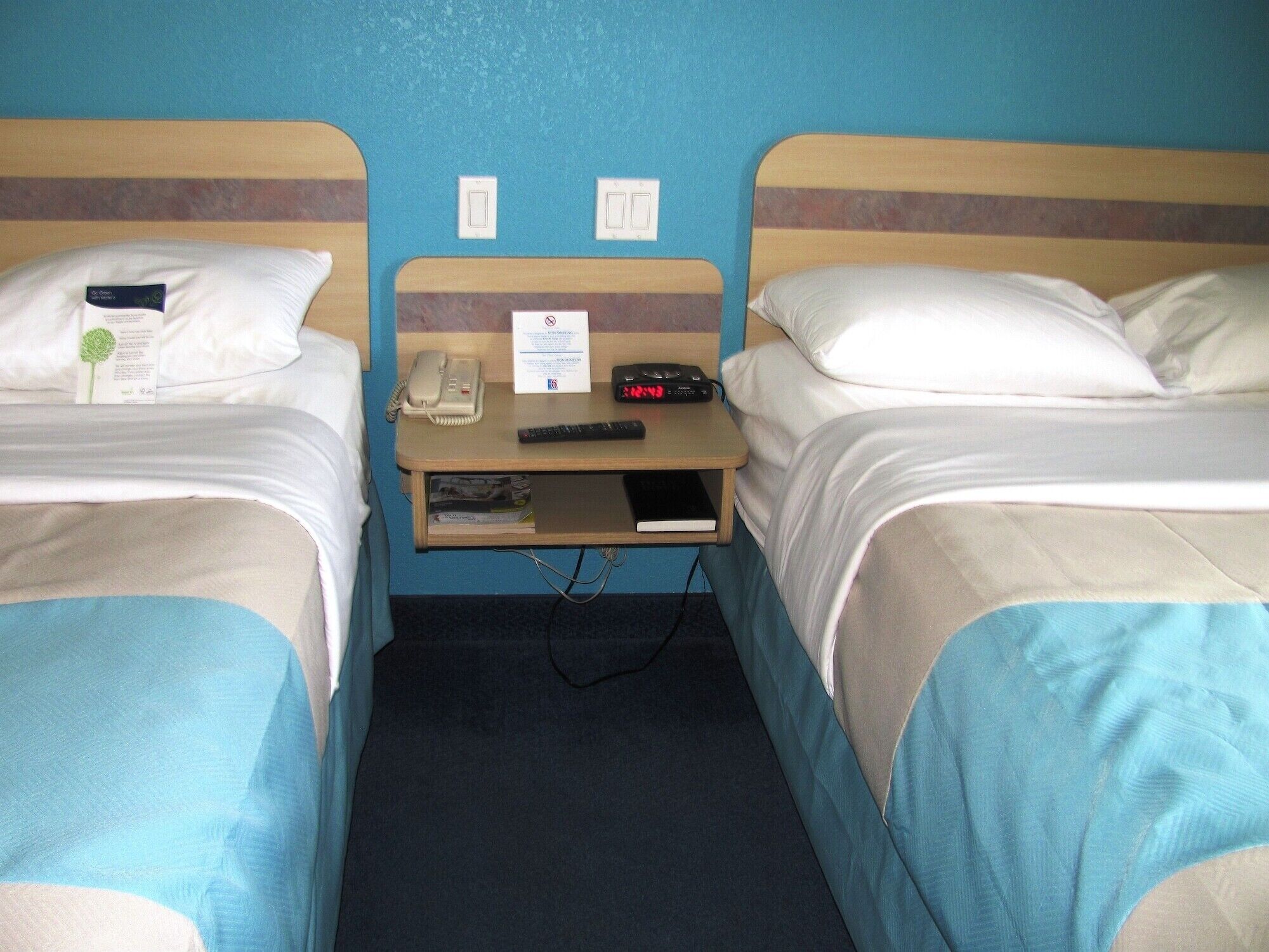 Een hotelkamer met twee eenpersoonsbedden, een nachtkastje met telefoon en klok, en een klein plankje met een boek.