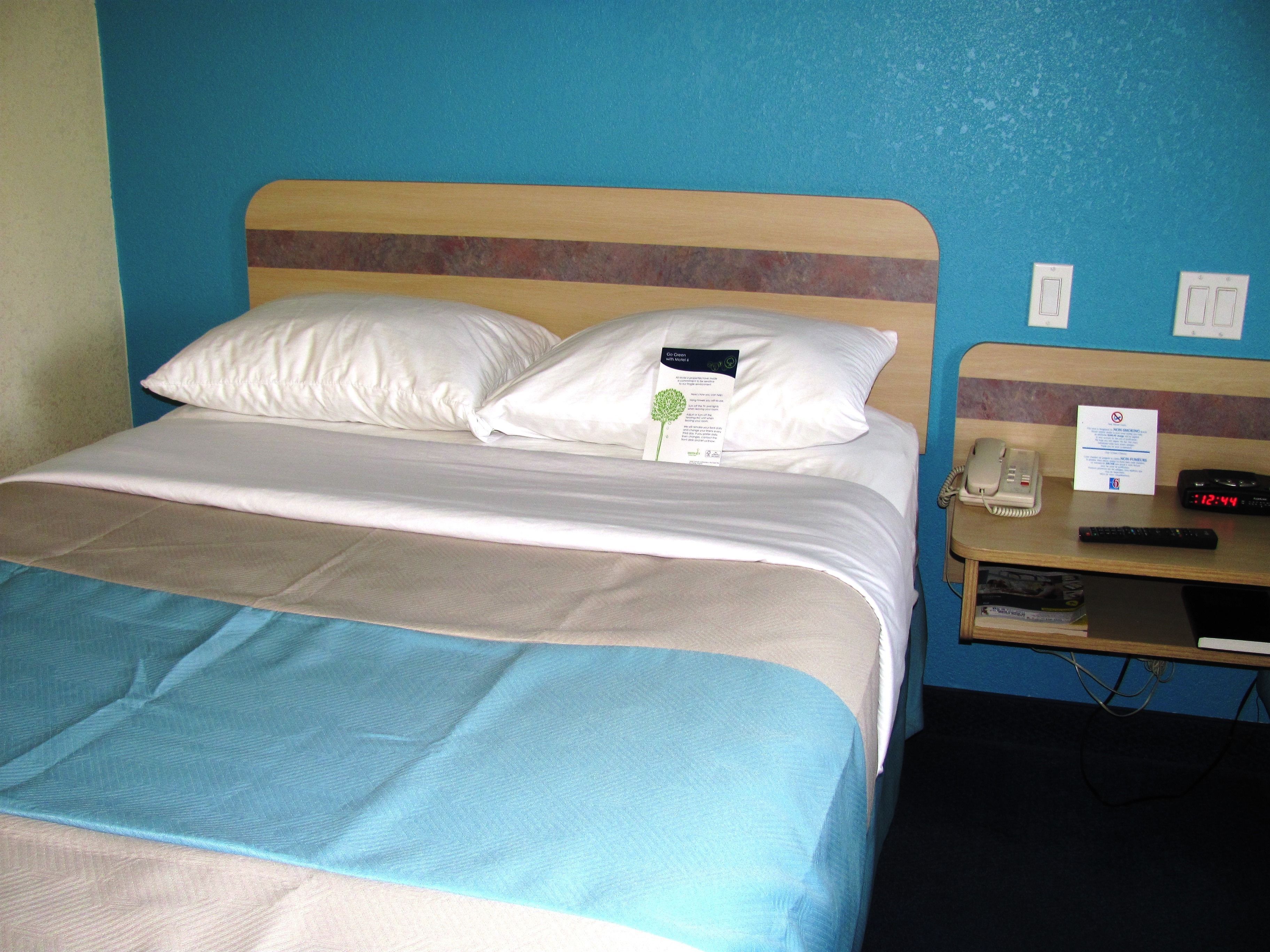 A hotel room with a bed, pillows, a bedside table with a phone, a remote control, and a sign on the wall.