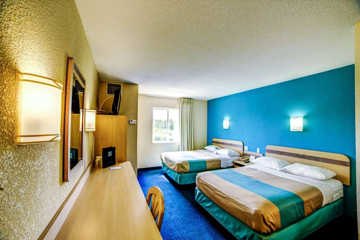 Hotellihuoneessa on kaksi sänkyä, työpöytä ja tuoli. Huoneessa on ikkuna, jossa on verhot, seinään kiinnitetty valaisin ja ovi.