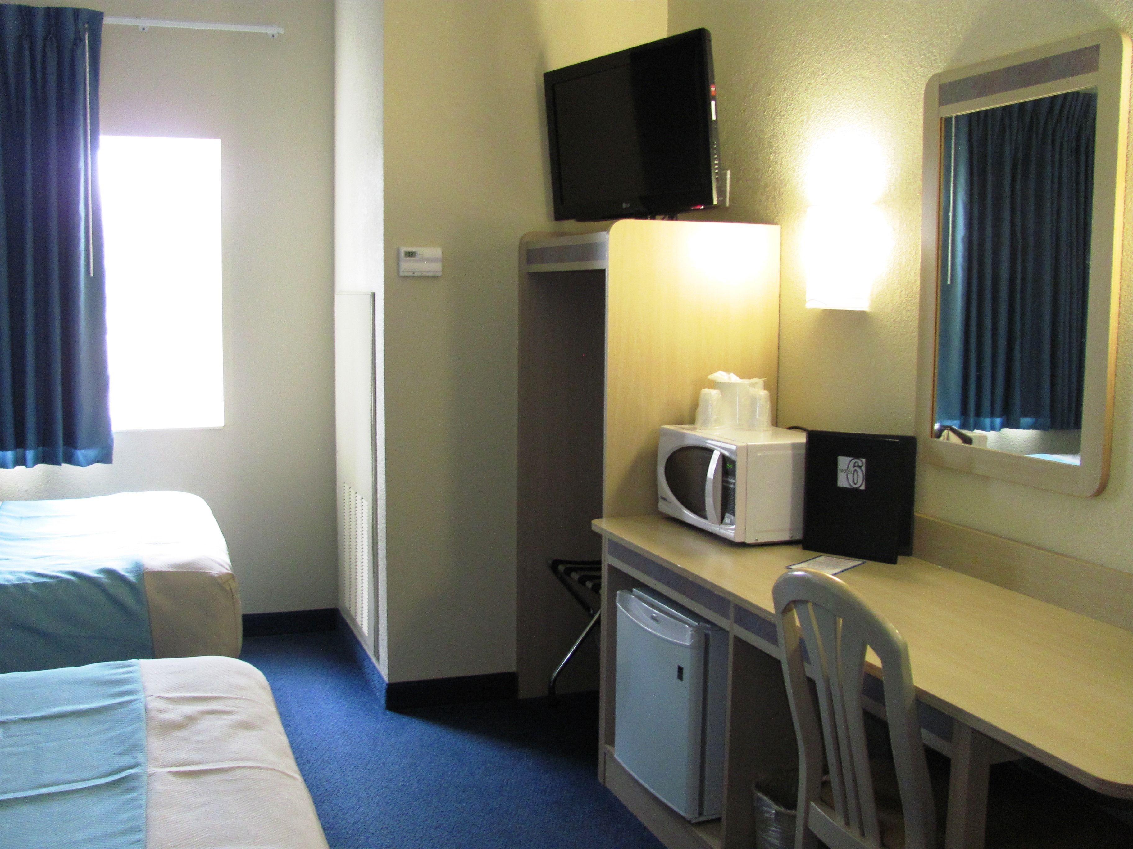 Quarto de hotel com cama, escrivaninha com micro-ondas, cadeira, pequeno frigorífico, televisão e janela com cortinas.