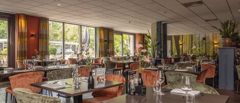 Un restaurante con varias mesas y sillas, ventanas amplias y plantas de interior.