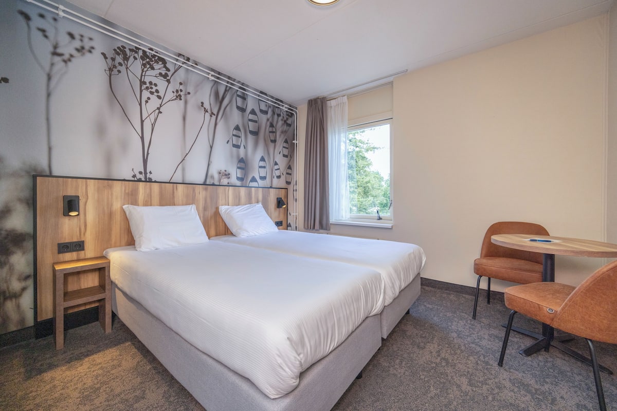 A hotel room with a large bed, a wooden nightstand, a small table with a chair, and a window with curtains.