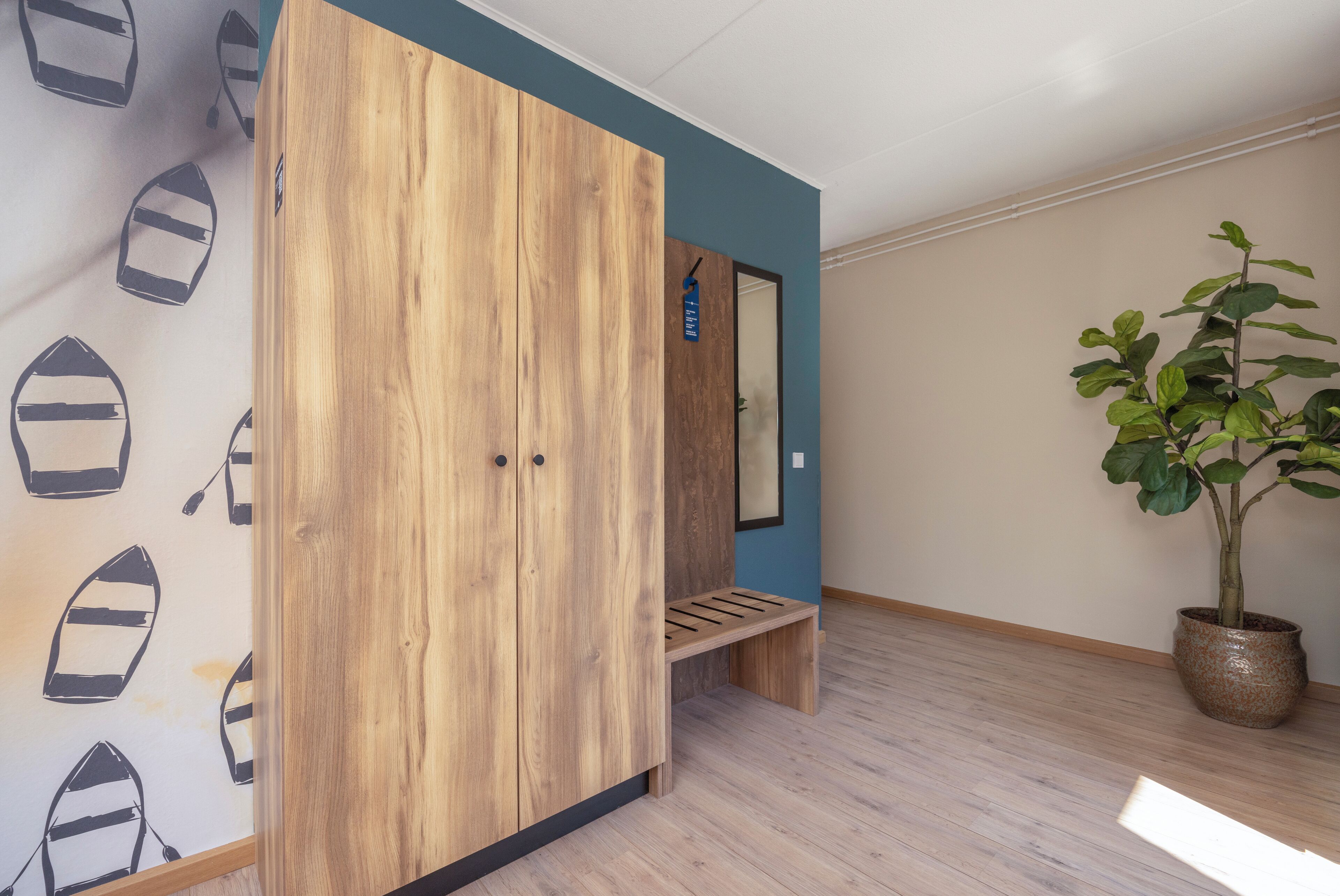 A room with a wooden wardrobe, a bench, a potted plant, and a wall with a boat pattern.