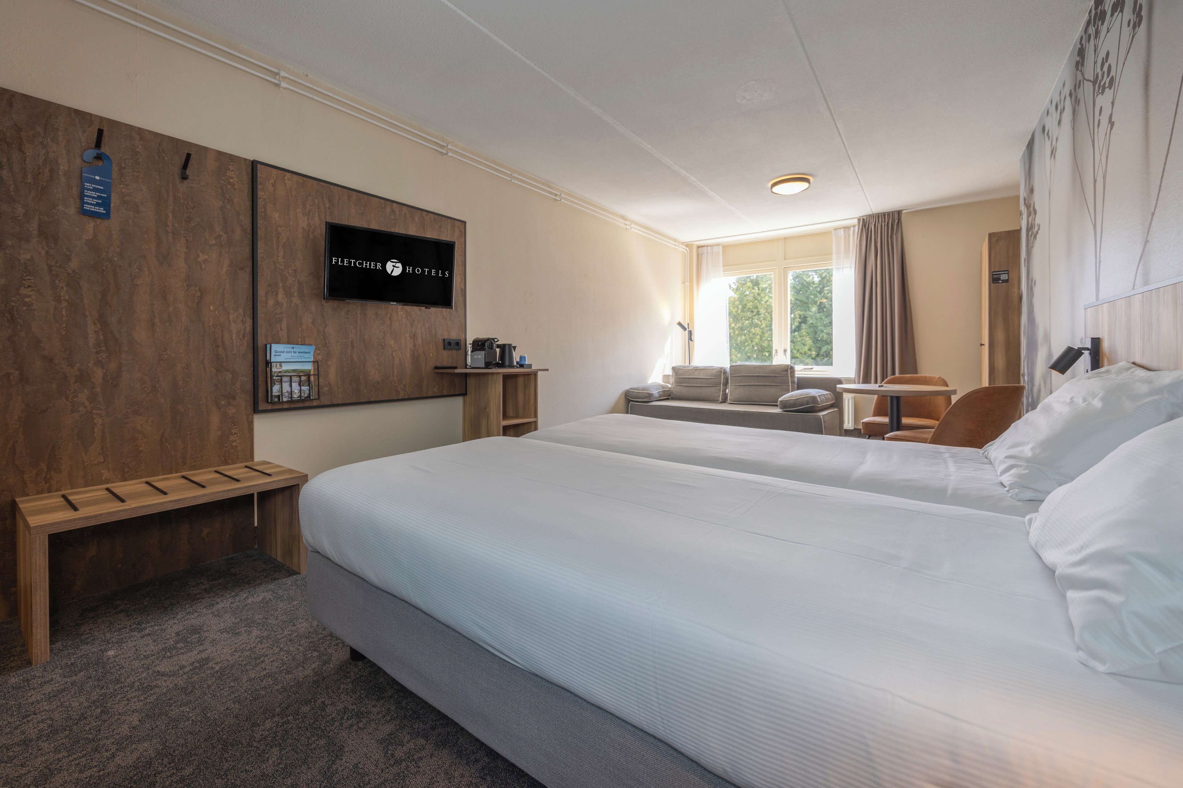 A hotel room with two beds, a wooden bench, a desk with a chair, a small table, and a window with curtains.