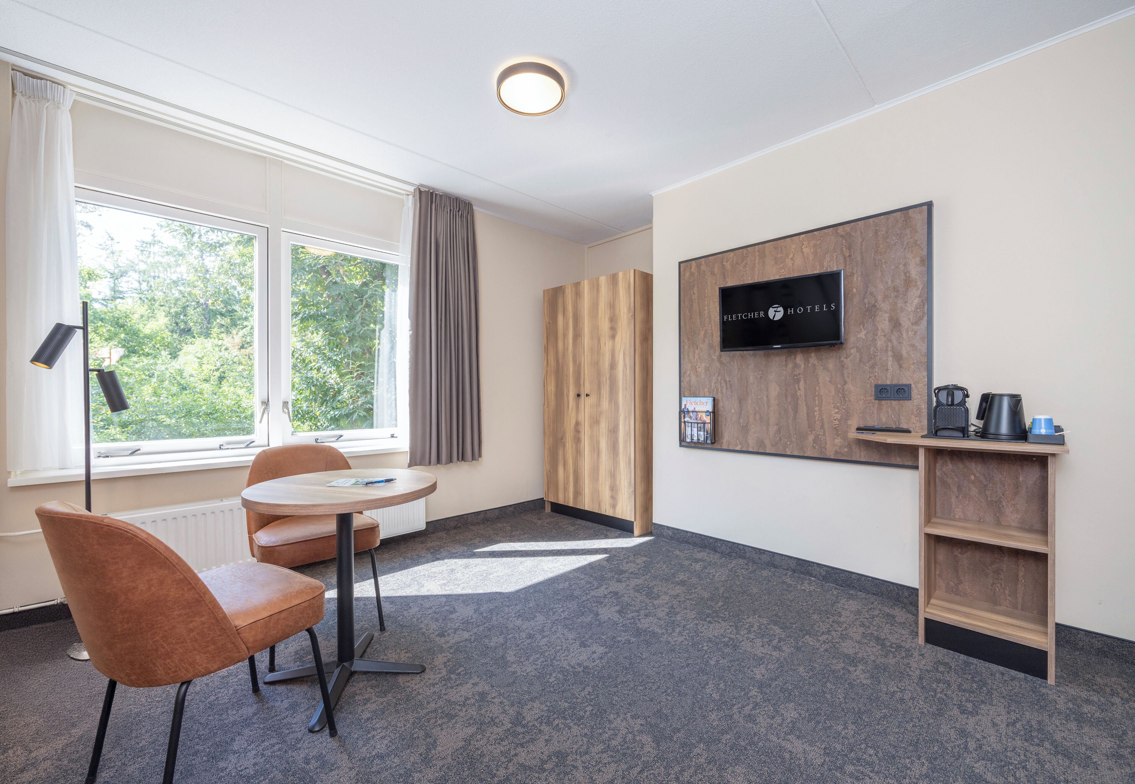 Een hotelkamer met een ronde tafel, twee stoelen, een televisie, een kledingkast en een koffiehoek.