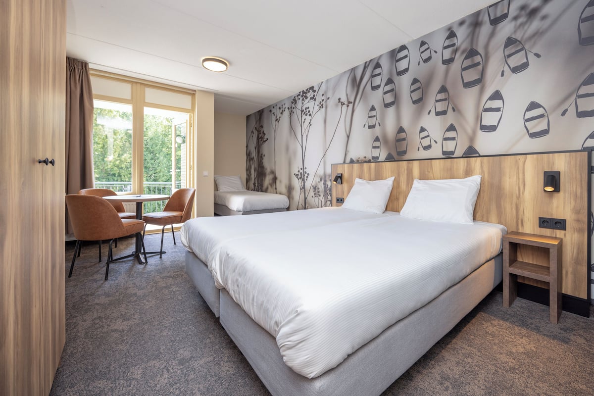 A hotel room with a large bed, a desk with a chair, a window with curtains, and a wall with a boat pattern.