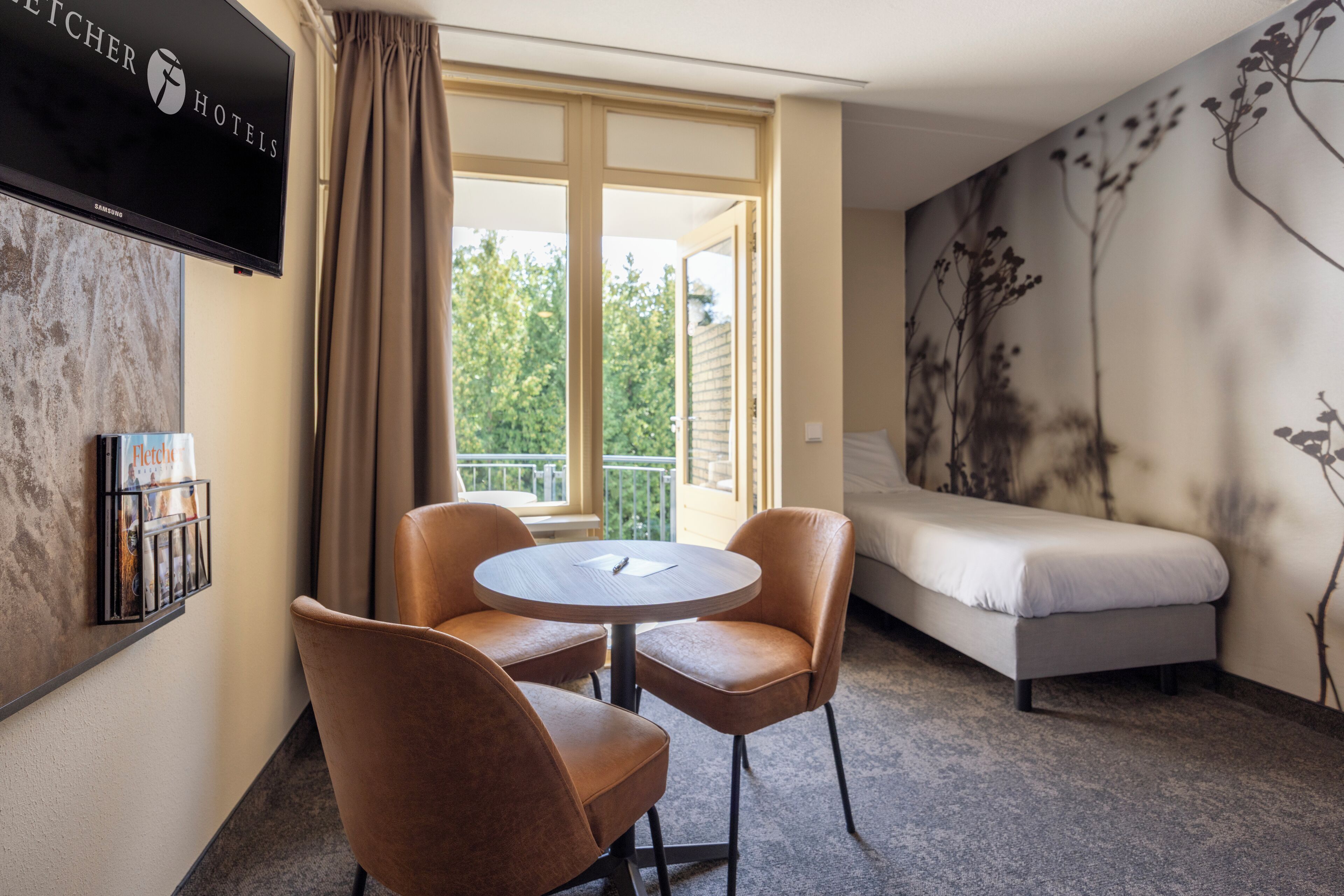 Ein Hotelzimmer mit einem Bett, einem runden Tisch mit zwei Stühlen, einem an der Wand befestigten Fernseher und einem Fenster mit Vorhängen.