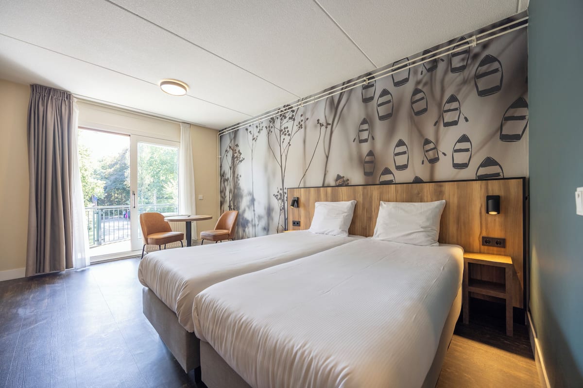 A hotel room with two beds, a wooden headboard, a small table, a chair, and a view of the outdoors through a large window.