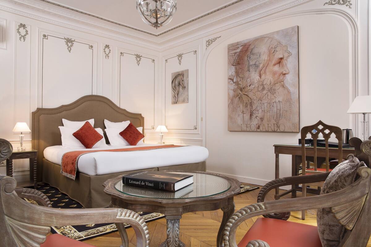 Una habitación de hotel con una cama grande, una mesa de centro de vidrio, una silla y un cuadro en la pared.