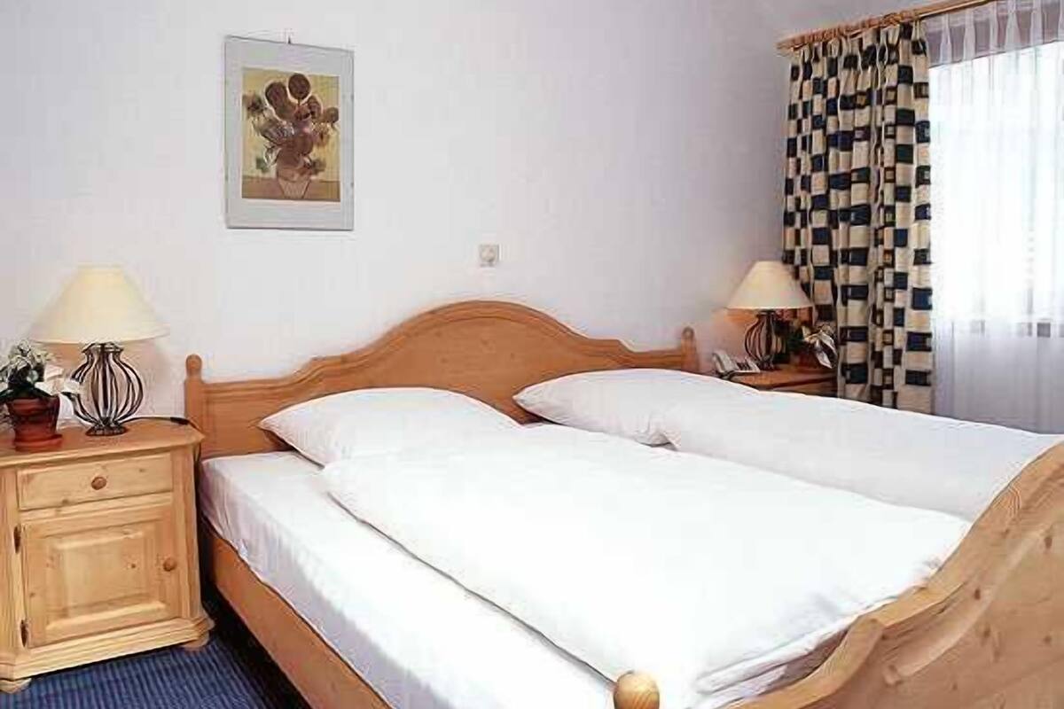 A double bed with white linens, a wooden headboard, two bedside tables with lamps, a framed picture on the wall, and checkered curtains on the window.