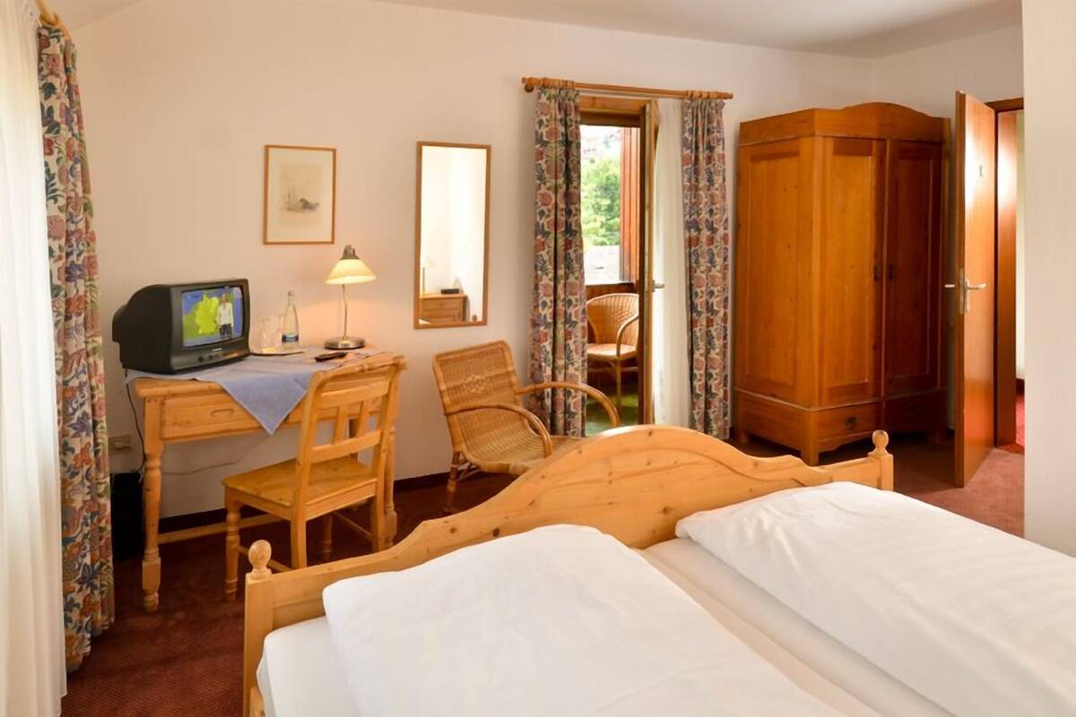 A hotel room with a wooden bed, a desk with a television, a chair, a wardrobe, and a window with curtains.