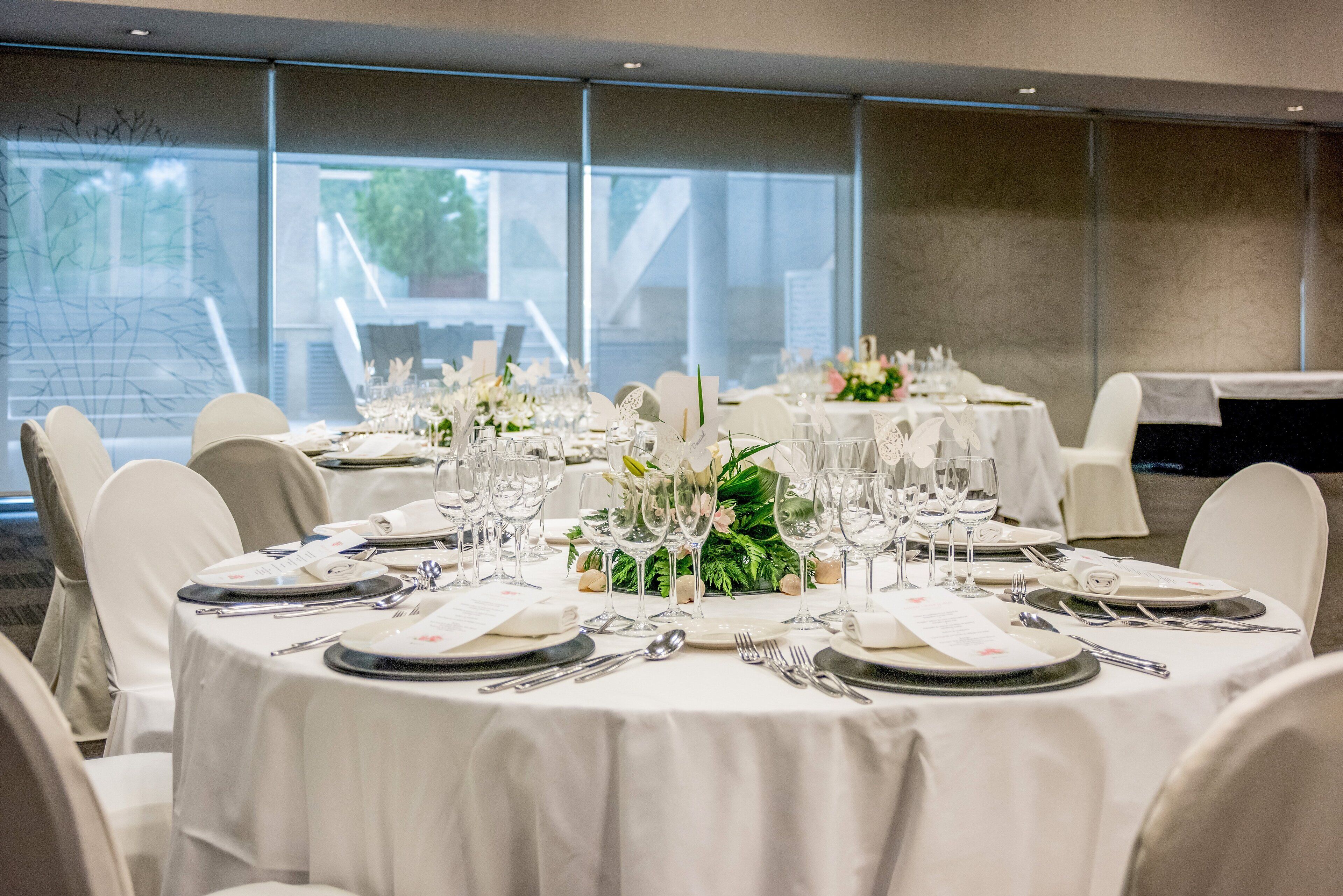 A banquet hall with round tables set for a formal event, featuring white tablecloths, floral centerpieces, and a view of a building through large windows.