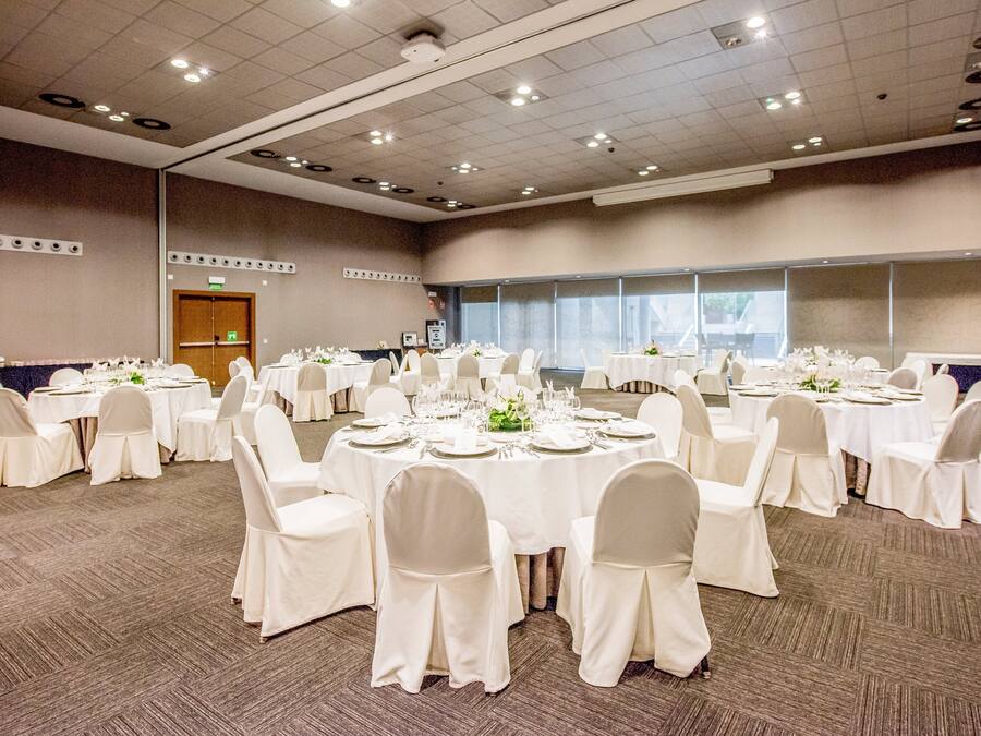Un salón de conferencias amplio con varias mesas redondas, cada una con mantel blanco, sillas y cubertería.