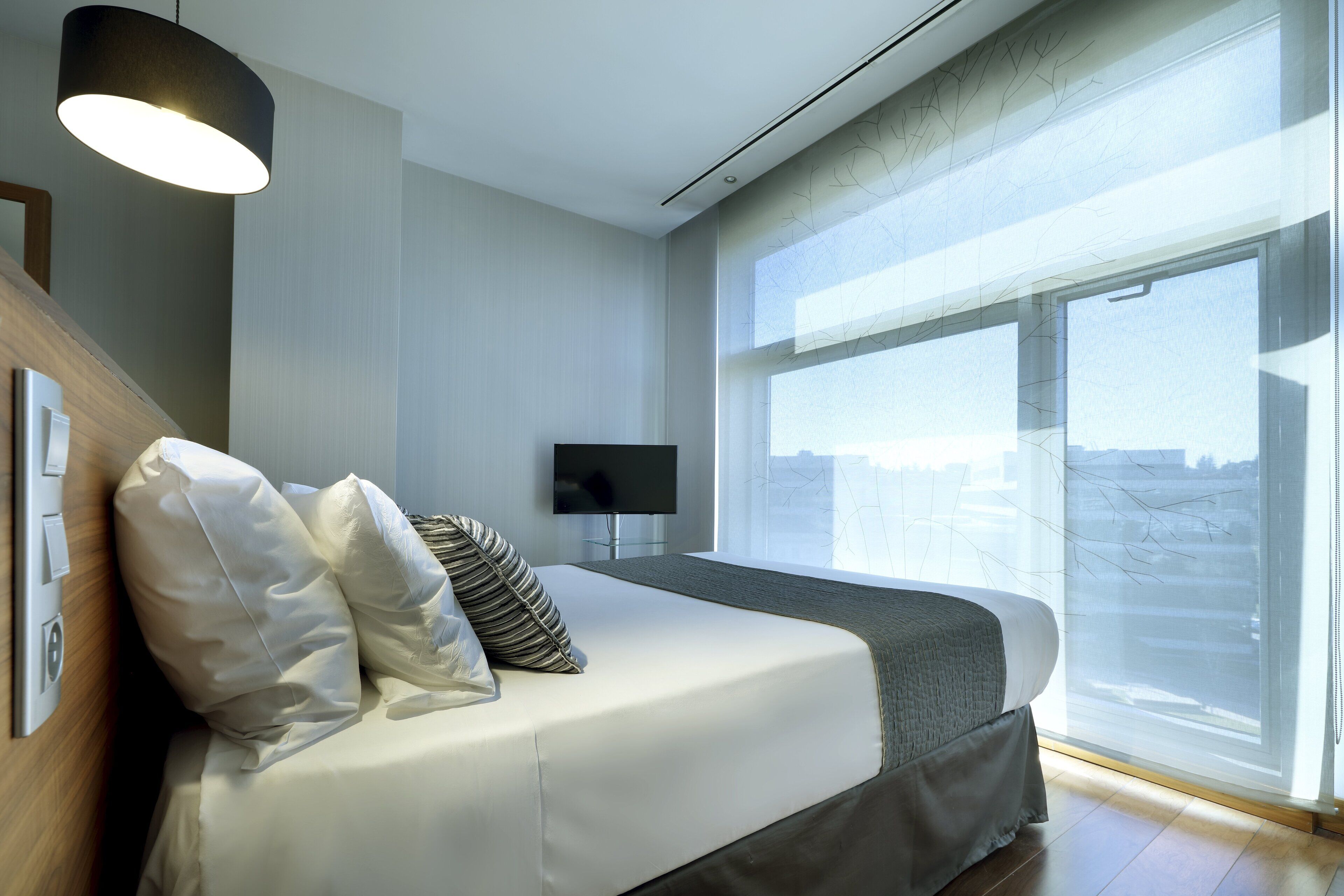 Una habitación de hotel moderna con una cama grande, un televisor de pantalla plana y un ventanal con vistas a la ciudad.