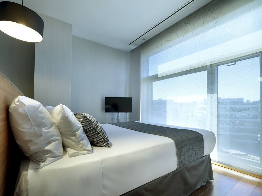 Una habitación de hotel moderna con una cama grande, un televisor de pantalla plana y un ventanal con vistas a la ciudad.