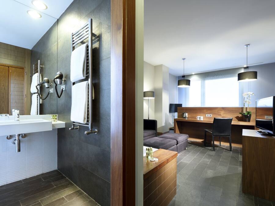 A modern bathroom with a large sink, towel rack, and a wooden vanity. The room features dark tiles, a wooden door, and recessed lighting.