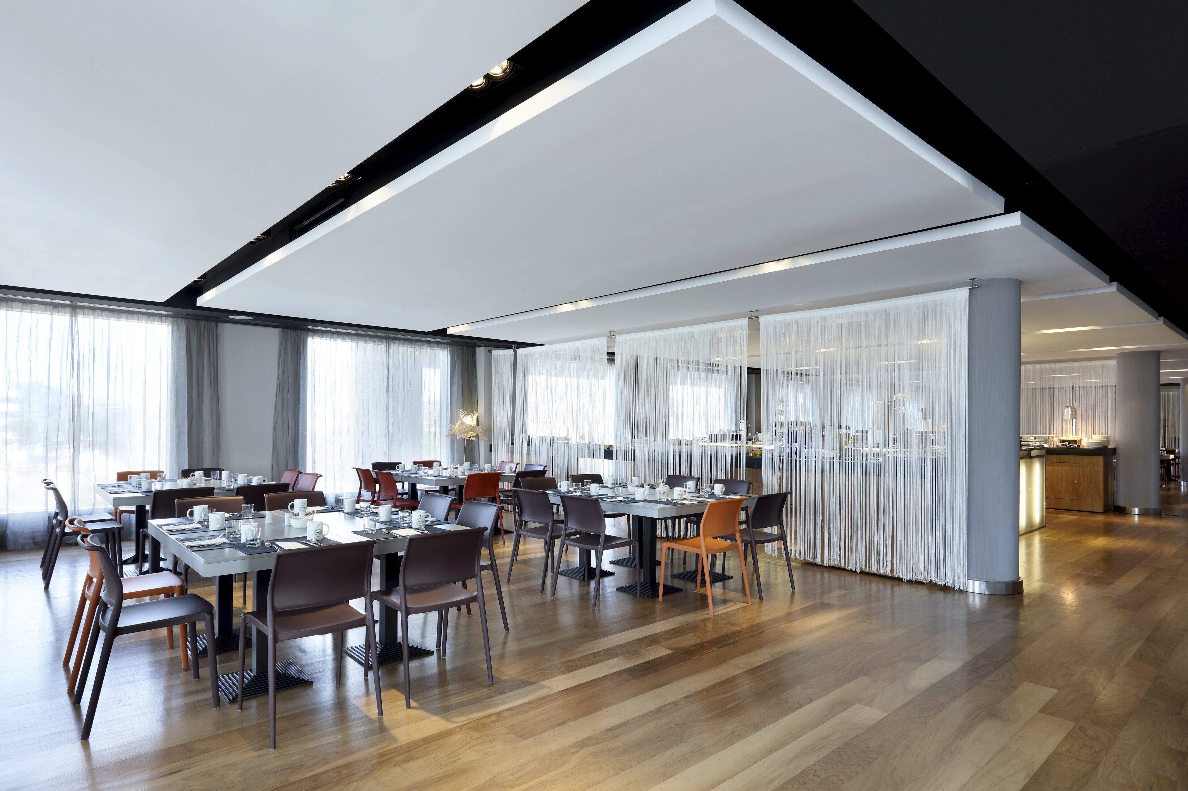 Un vaste espace dédié à la salle à manger, avec de nombreuses tables et chaises, de grandes fenêtres agrémentées de rideaux et un design moderne au niveau du plafond.
