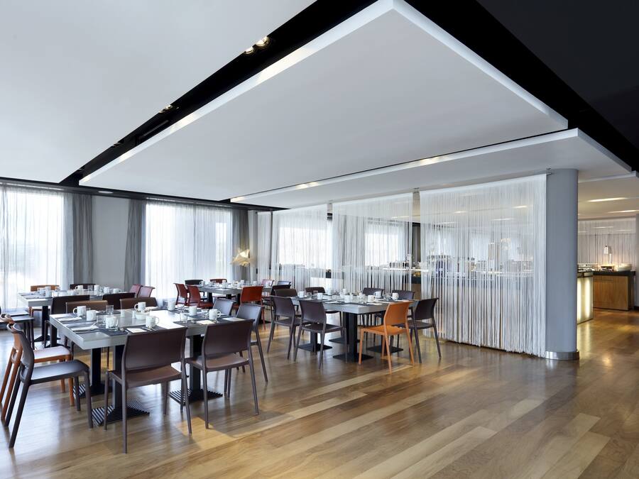 Un vaste espace dédié à la salle à manger, avec de nombreuses tables et chaises, de grandes fenêtres agrémentées de rideaux et un design moderne au niveau du plafond.