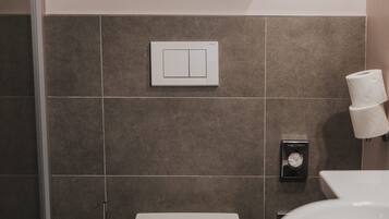 A modern bathroom with a toilet, a wooden shelf above the toilet, and a wall-mounted toilet paper holder.
